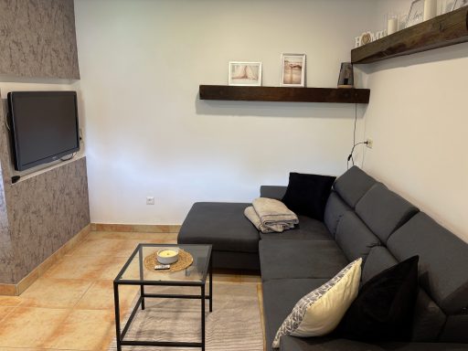 Wohnzimmer mit grauem Sofa, Glastisch und TV an der Wand. Deco-Elemente auf dem Regal.